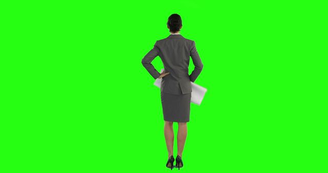 Businesswoman in a formal suit stands with her back towards the viewer, holding papers in her hand against a vibrant green screen background. This image is useful for corporate presentations, business training materials, or visual effects where a professional setting is incorporated. Ideal for graphic designers and video editors needing a versatile and customizable image.