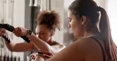 Female workout partners checking wrist devices while pulling cable handles in sunlit gym