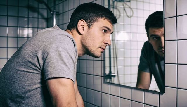 Young man leaning over bathroom sink studying reflection in mirror moody introspective