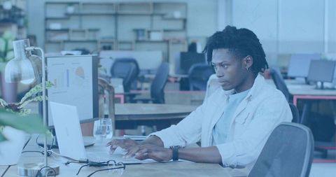 Focused man with dreads typing on laptop in open-plan coworking space with data charts