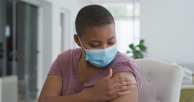 Woman wearing a mask examining a bandage on her arm while sitting indoors. Ideal for themes like vaccination, healthcare, safety, public health campaigns, and personal protection. This image can be used to promote vaccination drives, healthcare services, and medical guidance materials.