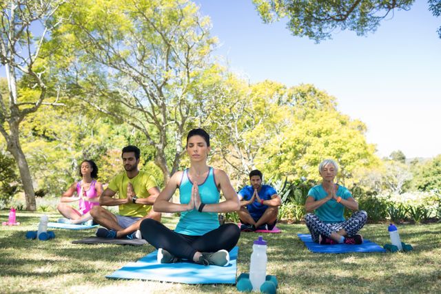 Group of people practicing yoga in a park on a sunny day. They are sitting on yoga mats in a meditative pose, surrounded by lush greenery. This image can be used for promoting outdoor fitness activities, wellness programs, and healthy lifestyle campaigns.