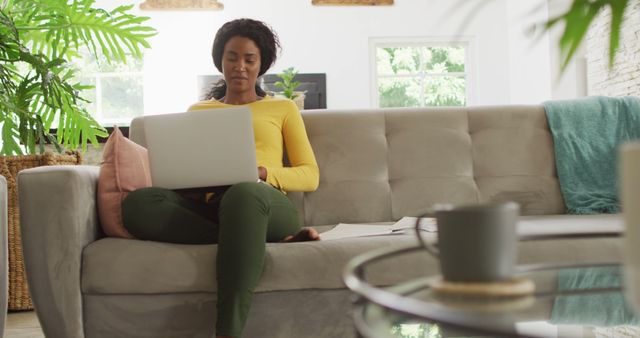 Young woman wearing yellow shirt sitting on sofa and working on laptop in a modern living room with green plants and minimalist decor. Perfect for content related to remote work, home office setup, productivity, work-life balance, modern lifestyle, and cozy home environments.