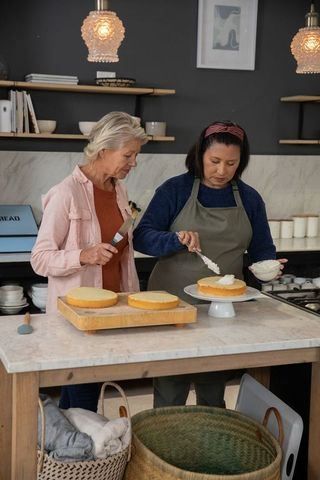 Mature women frosting cake layers in cozy kitchen, teaching baking techniques and teamwork
