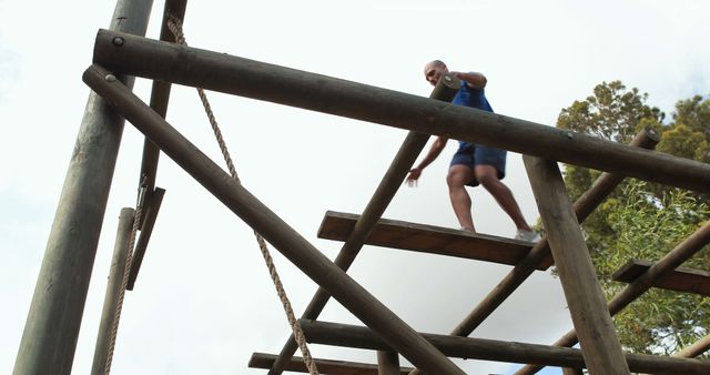 Man participating in an outdoor obstacle course, balancing on wooden beams and ropes on a clear, sunny day. Ideal for illustrating fitness activities, outdoor adventures, training programs, and team-building events.