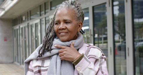 Senior african american woman smiling while touching scarf and checked coat on city sidewalk