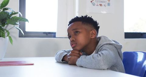 African American Boy Contemplating at Classroom Table, Gazing Toward Sunlit Window