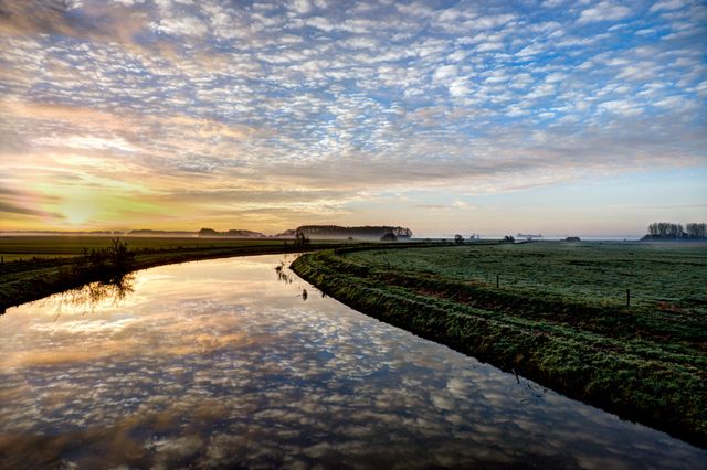 Beautiful sunrise scene over a calm river in a peaceful countryside setting. The sky is reflected perfectly in the placid water, creating a mirror-like effect. Lush green fields stretch out to the horizon with a silhouette of distant trees, adding to the serene ambiance. Ideal for use in nature-themed projects, relaxation visuals, travel brochures, and environmental conservation materials.