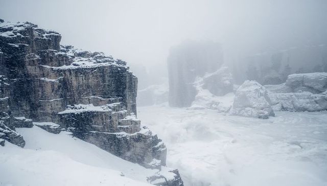 Towering stratified cliff shedding snow into frozen alpine gorge during blizzard