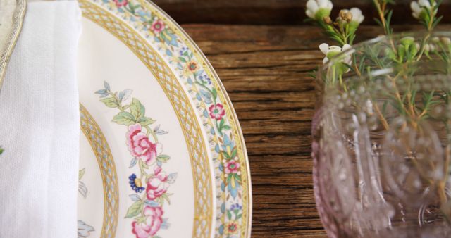 A close-up of an elegant table setting features a vintage plate with a floral design, a neatly folded napkin, and a sophisticated pink glass placed on a rustic wooden table. This image is ideal for use in articles or advertisements related to fine dining, table decoration ideas, event planning, and lifestyle blogs promoting luxury and vintage aesthetics.