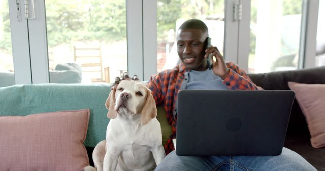 African American man multitasking while working from home, sitting on sofa with smartphone and laptop. Pet dog accompanying him, creating a casual, relatable home office scene. Useful for themes around remote work, pet companionship, work-life balance, and modern lifestyle.