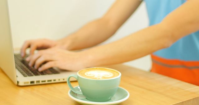 Man working on a laptop. Coffee cup with latte art is in the foreground. Ideal for depicting themes of remote work, productivity, coffee culture, and working from a cafe or home office.