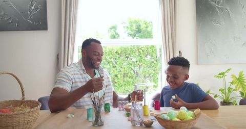 African american father and son painting easter eggs together at sunlit kitchen table