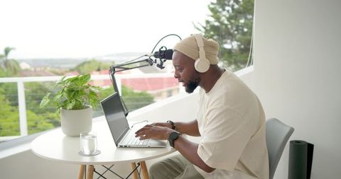 Recording podcast on balcony: host wearing beanie and headphones typing on laptop