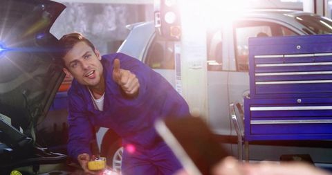 Mechanic giving thumbs up while checking car engine with smartphone and diagnostic tool