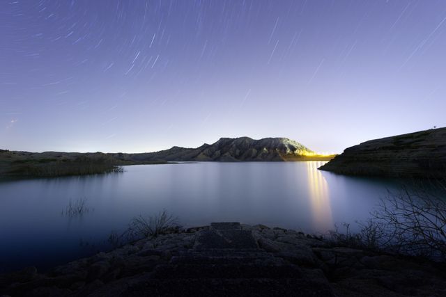 Tranquil night scene with star trails over a calm lake reflecting a mountain range. Perfect for projects involving nature, serenity, evening, or peaceful landscapes. Ideal for backgrounds, travel articles, and meditation brochures.