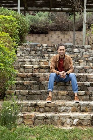 Relaxed man sitting on rustic stone steps in garden under pergola, casual autumn outfit