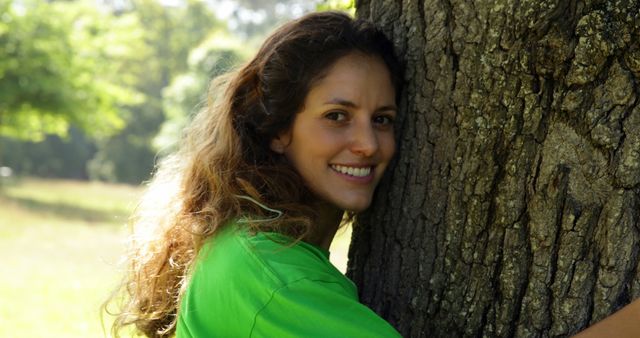 Young woman hugging tree in sunny park. She wears a casual green shirt, symbolizing an eco-friendly lifestyle. Ideal for use in environmental campaigns, nature conservation material, wellness promotions, or illustrations of happiness and leisure in outdoor settings.