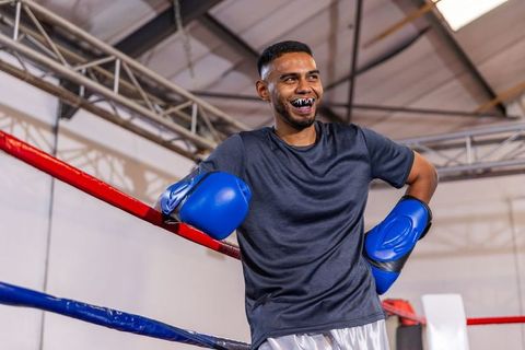Smiling male boxer leaning on ropes wearing blue gloves and black mouthguard warehouse gym