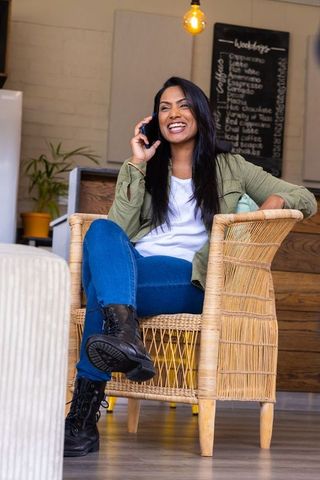 Asian woman laughing while talking on smartphone in cozy cafe lounge with wicker chair