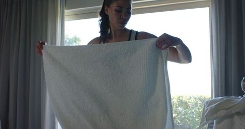 Backlit woman holding and stretching white towel in sunlit minimalist bedroom by window