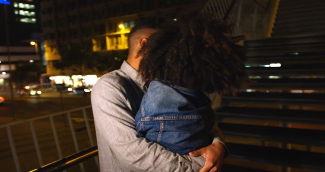 Father holding and hugging his young child in a city setting at night under streetlights. The image captures a tender moment of affection and bonding between parent and child amidst the urban environment. This could be used in advertisements, articles, and campaigns focusing on family values, parenthood, or nighttime urban environments.