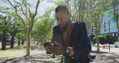 Business commuter checking smartphone while leaning on bicycle in sunlit urban parkway