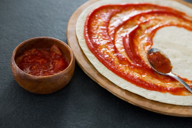 Image shows pizza dough being spread with tomato sauce using a spoon. A wooden bowl filled with tomato sauce is placed next to the dough. This image is perfect for use in cooking blogs, recipe websites, culinary magazines, and food-related advertisements.