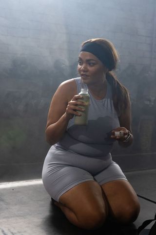 Kneeling african american woman holding shaker drinking post-workout in dim gym studio