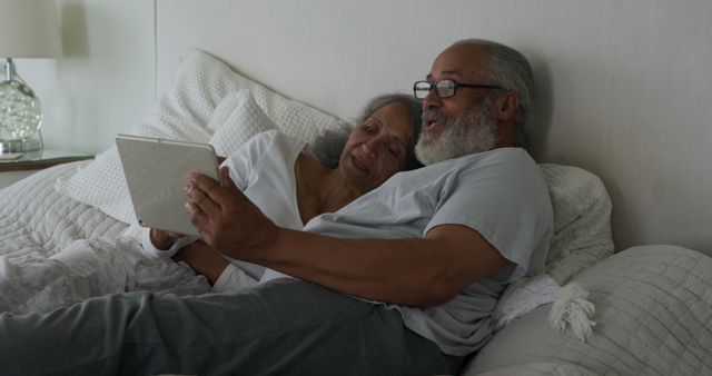 Senior biracial couple relaxing in bed at home. They are sharing a moment of joy while looking at a tablet.