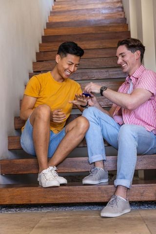 Asian and caucasian friends laughing and testing gadget while sitting on wooden stairs