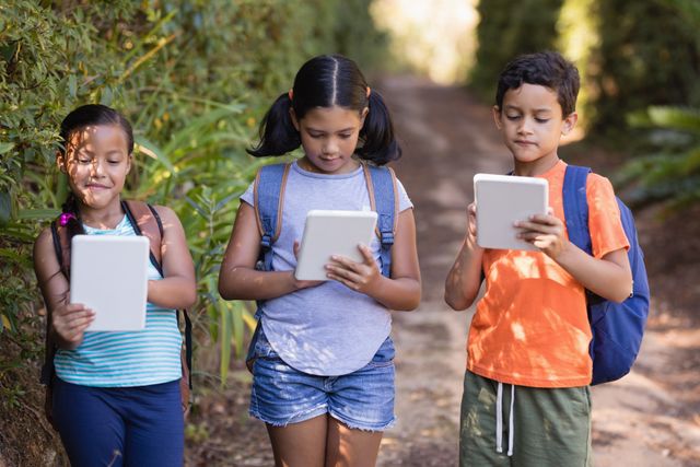 Three children are walking in a natural park while using digital tablets. They are engaged with their devices, indicating a blend of technology and outdoor activity. This image can be used for educational content, technology in nature, outdoor learning, or promoting digital literacy among children.
