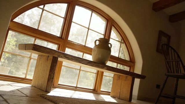 Sunlit reclaimed wooden bench with ceramic jug at arched window, cozy farmhouse nook