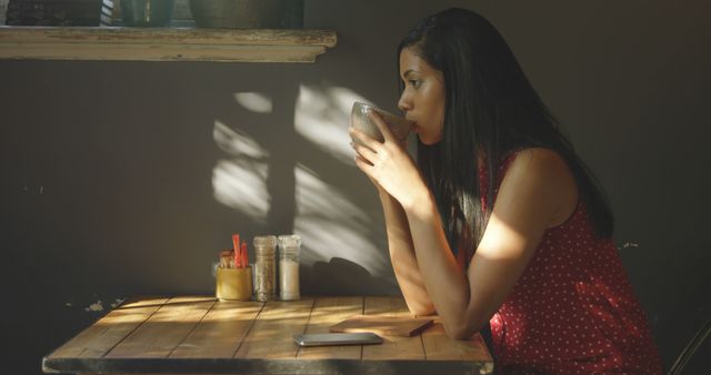 Woman enjoying coffee alone near a sunlit wooden table in a cozy cafe. Perfect for illustrating leisure and relaxation, coffee shop culture, moments of solitude, and thoughtful contemplation. Useful for articles, blog posts, advertisements for cafes, and lifestyle promotions.