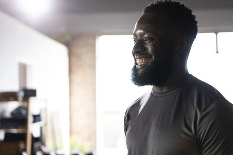 Smiling african american man standing in sunlit loft wearing gray tee and full beard