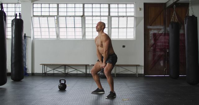 Muscular man squatting while lifting kettlebell in gym. Ideal for fitness blogs, workout guides, strength training programs, exercise technique demonstrations, and athleticism promotion.