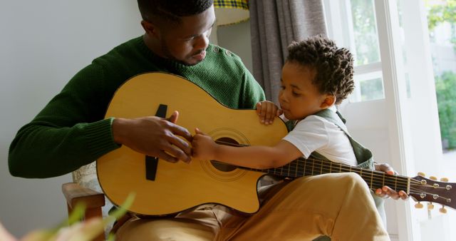 Father teaching young son to play guitar inside home during daylight, bonding over music. Ideal for content related to parenting, music education, family life, and promoting positive family interactions. Suitable for blogs, parenting articles, educational materials, and lifestyle magazines.