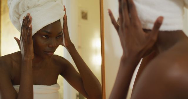 African american woman with towel touching her face and watching in mirror in bathroom. health and beauty concept.
