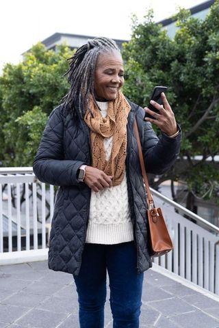 Senior african american woman smiling and checking smartphone on urban walkway with knit scarf