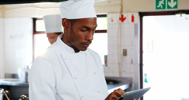 Chef wearing white uniform and hat is using tablet in modern kitchen for recipe management and menu planning. Ideal for content related to culinary arts, technology in kitchens, professional chefs at work, modern cooking techniques, restaurant management, and digital tools in the culinary field.