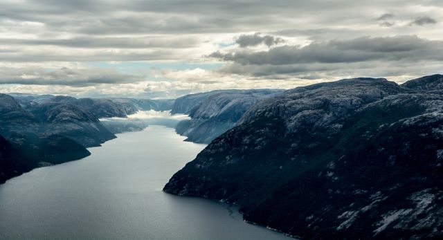 This breathtaking view showcases a tranquil fjord flanked by rugged mountains under an overcast sky, offering a dramatic and serene natural setting. Ideal for use in travel magazines, outdoor adventure adverts, environmental awareness campaigns, and landscape photography collections.