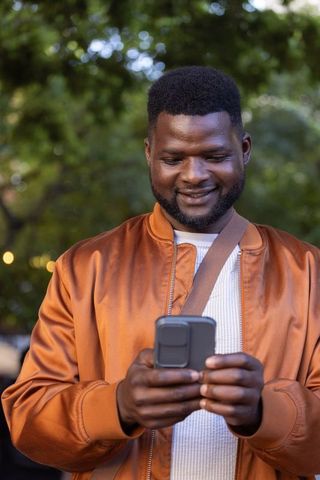 African american man smiling while texting on smartphone in park at golden hour