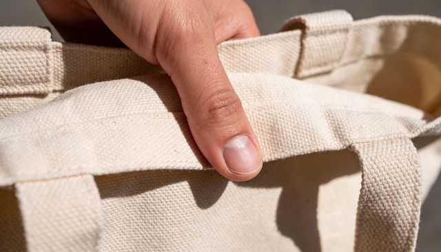 Holding canvas tote strap with adult right hand pressing beige woven fabric texture closeup