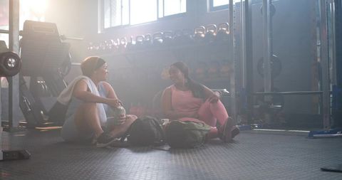 Diverse women taking gym break, chatting on rubber floor in sunlit fitness studio