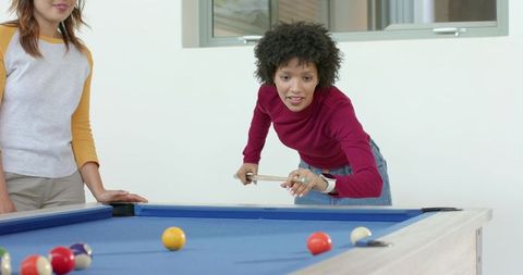 Young multiracial women playing pool and aiming cue on bright blue-felt game table