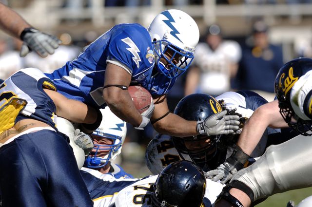 Football players appear engaged in a fast-paced, competitive game. The players in blue uniforms are tackling one in black to prevent progress. Perfect for illustrating the intensity and dynamics of American football, sports-related articles, or promotions emphasizing teamwork and athleticism.