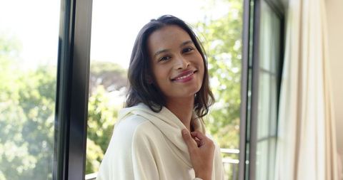 Young woman smiling on balcony wearing cozy hoodie near sunlit sliding door