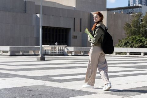 Redhead student walking across brutalist urban plaza carrying backpack and green folder
