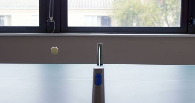 This depicts a ping pong ball captured in mid-air as it travels over the table tennis net, with a large window in the background. Ideal for use in subjects related to sports, recreation, indoor activities, and hobbies. Useful for websites or articles about ping pong, table tennis, indoor gaming spaces, leisure activities, and competitive sports.