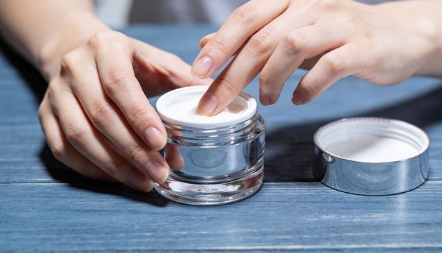 Dipping finger into white cream jar on blue tabletop for moisturizing skin care routine